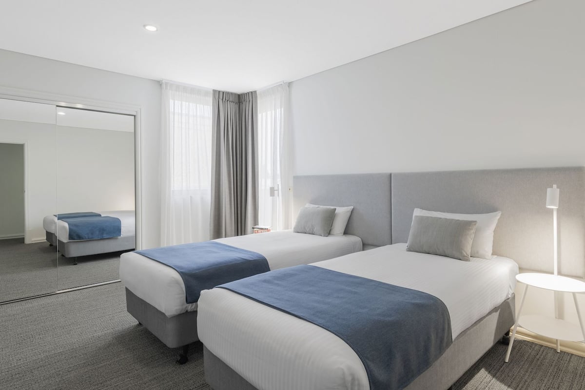 A modern hotel room with two beds, a large mirror, and a bedside table with a lamp.