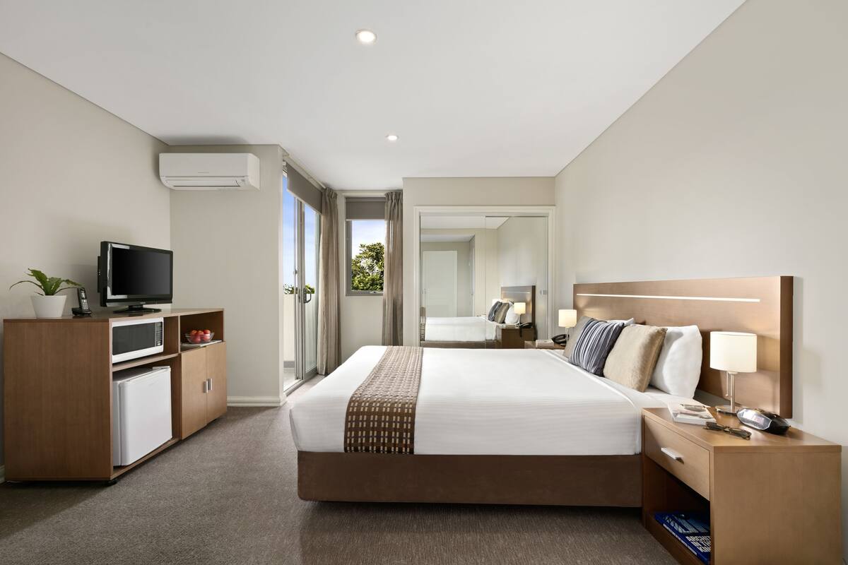 A modern hotel room with a bed, bedside table, TV, mini-fridge, and a view of the outdoors.