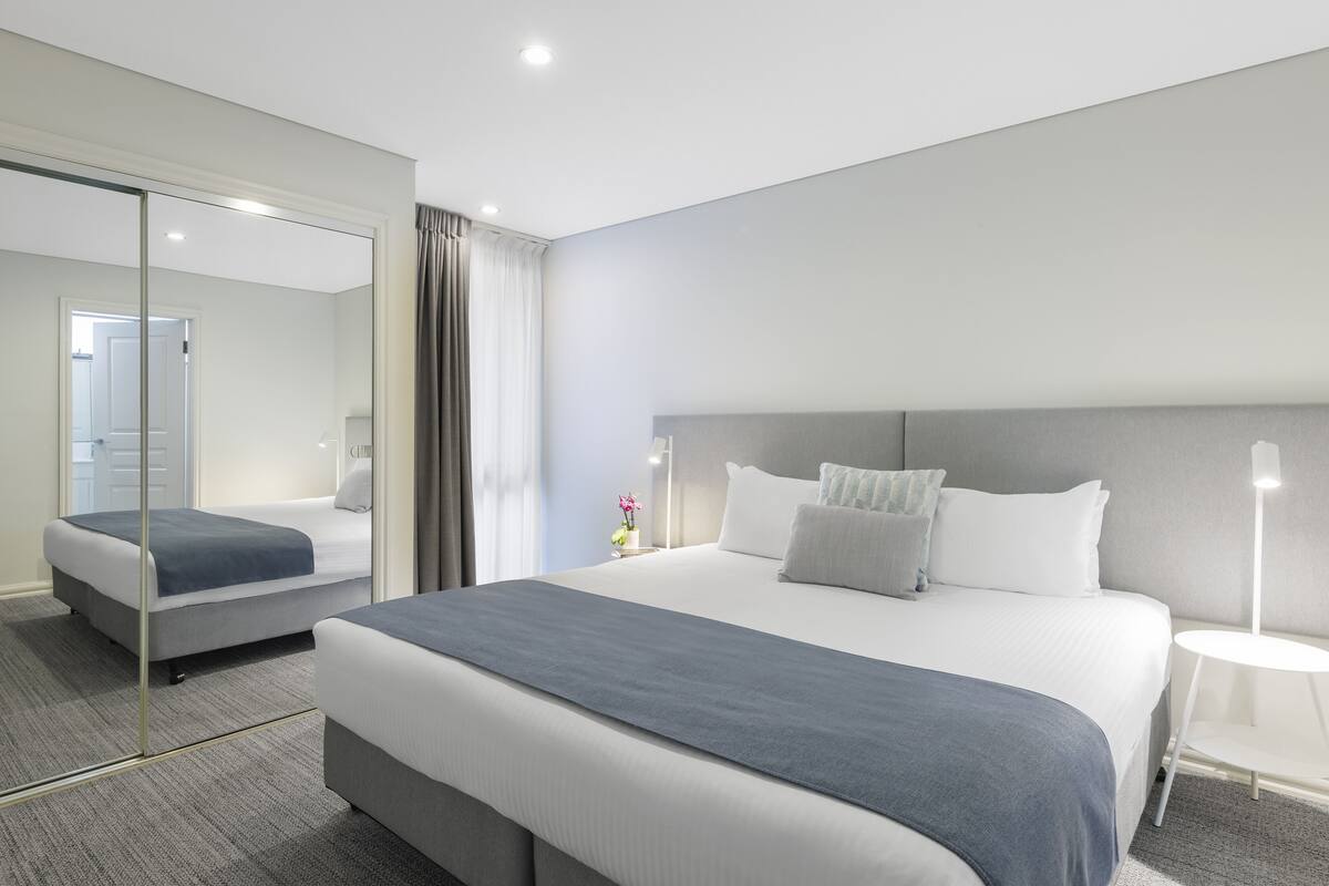 A modern hotel room with a large bed, a bedside table, a mirror, and a view of the hallway.