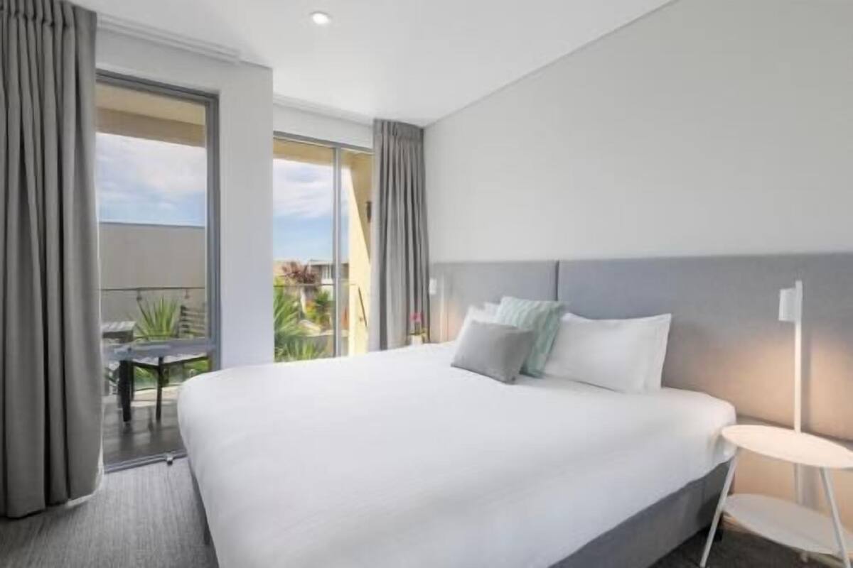 A hotel room with a large bed, a bedside table, a window with curtains, and a view of the outdoors.