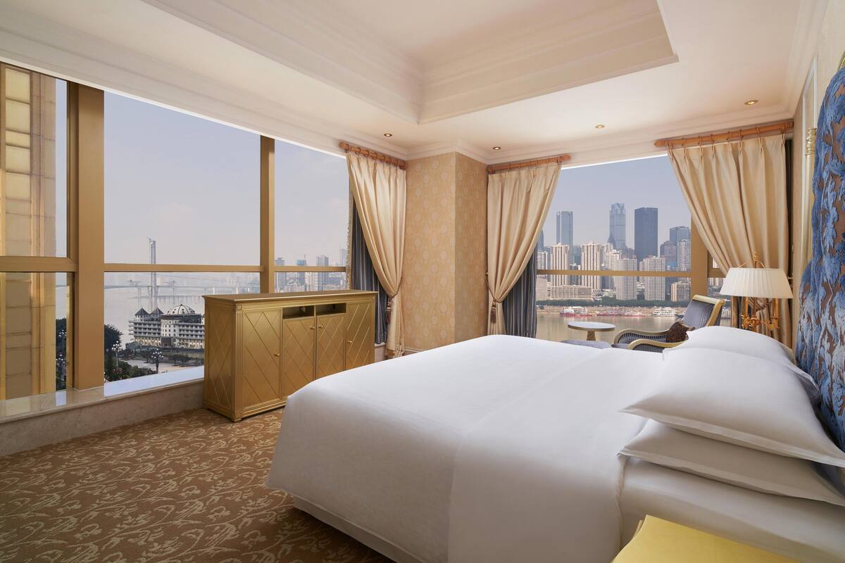 A hotel room with a large bed, a wooden wardrobe, a small table, and a chair. The room has large windows with curtains, offering a view of a city skyline.