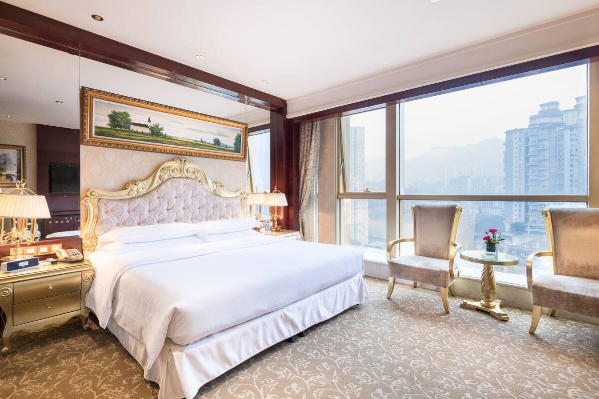 A hotel room with a large bed, two armchairs, a small table, and a large window with a city view.