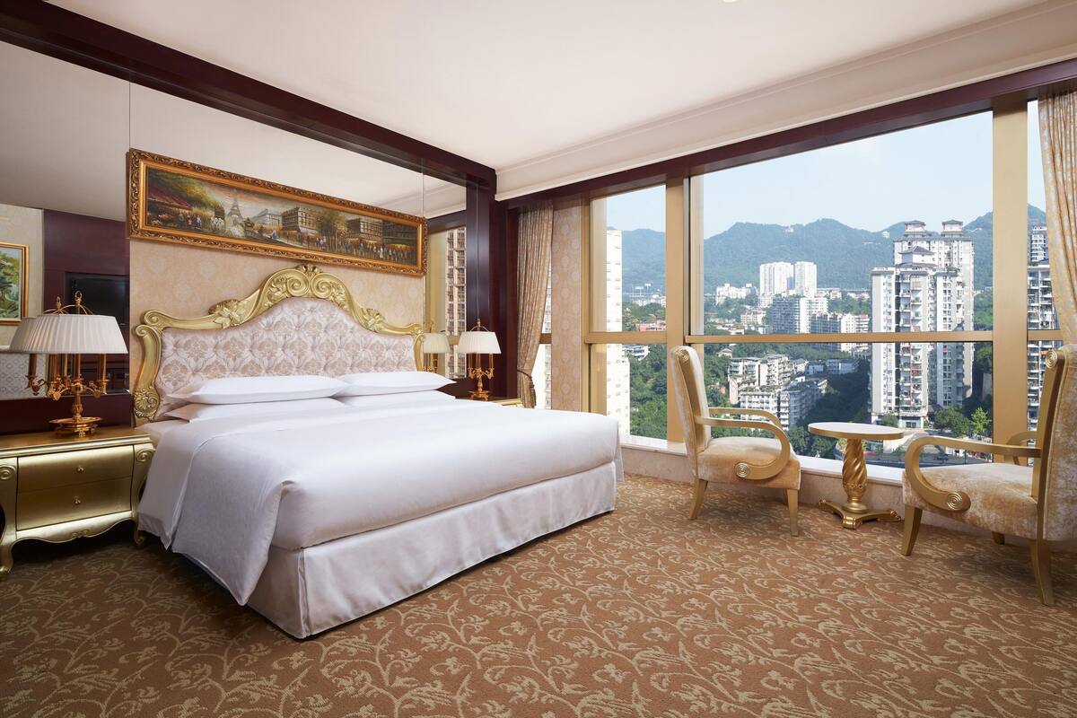 A hotel room with a large bed, a nightstand with a lamp, a chair, a small table, and a large window offering a city view.