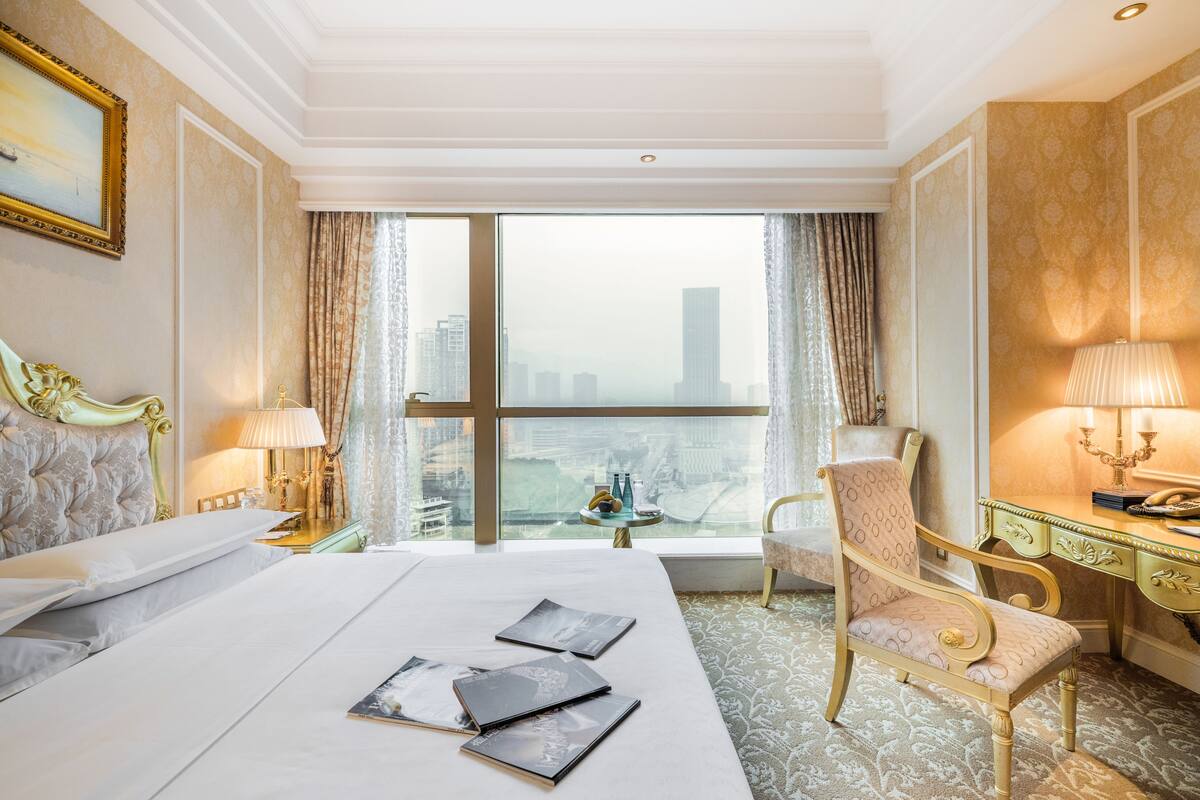 A hotel room with a large bed, a desk, a chair, and a view of the cityscape through the window.