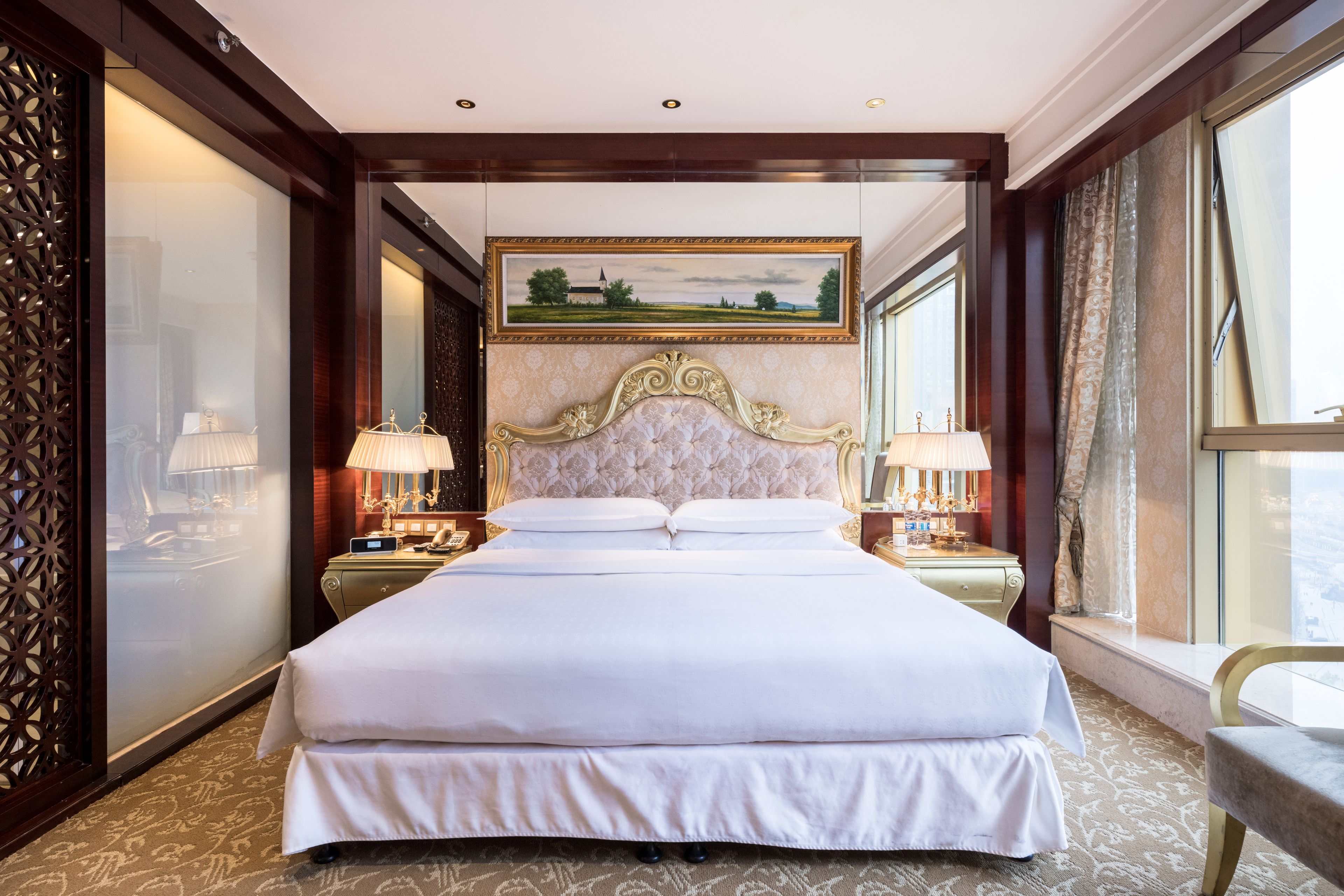 A large, centrally positioned bed with a decorative headboard, flanked by two bedside tables with lamps, in a room with patterned carpet and large windows.
