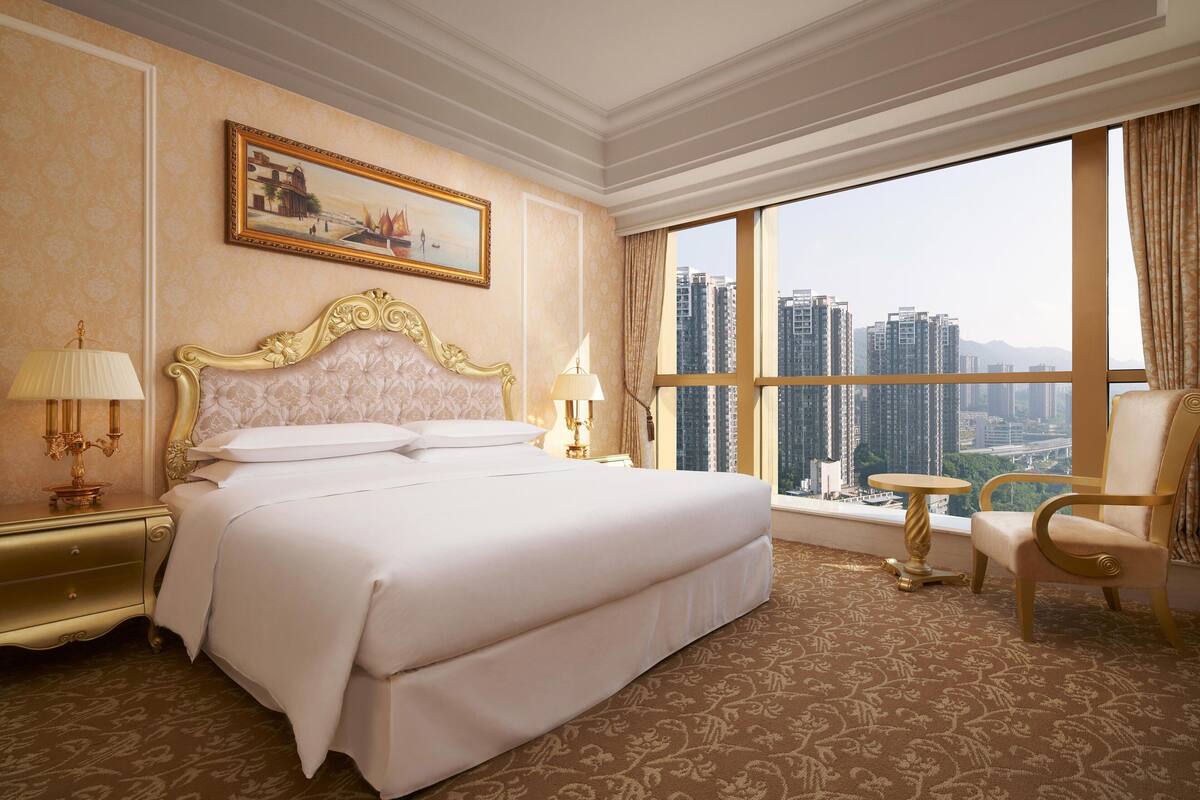 A hotel room with a large bed, a chair, a small table, and a view of high-rise buildings.