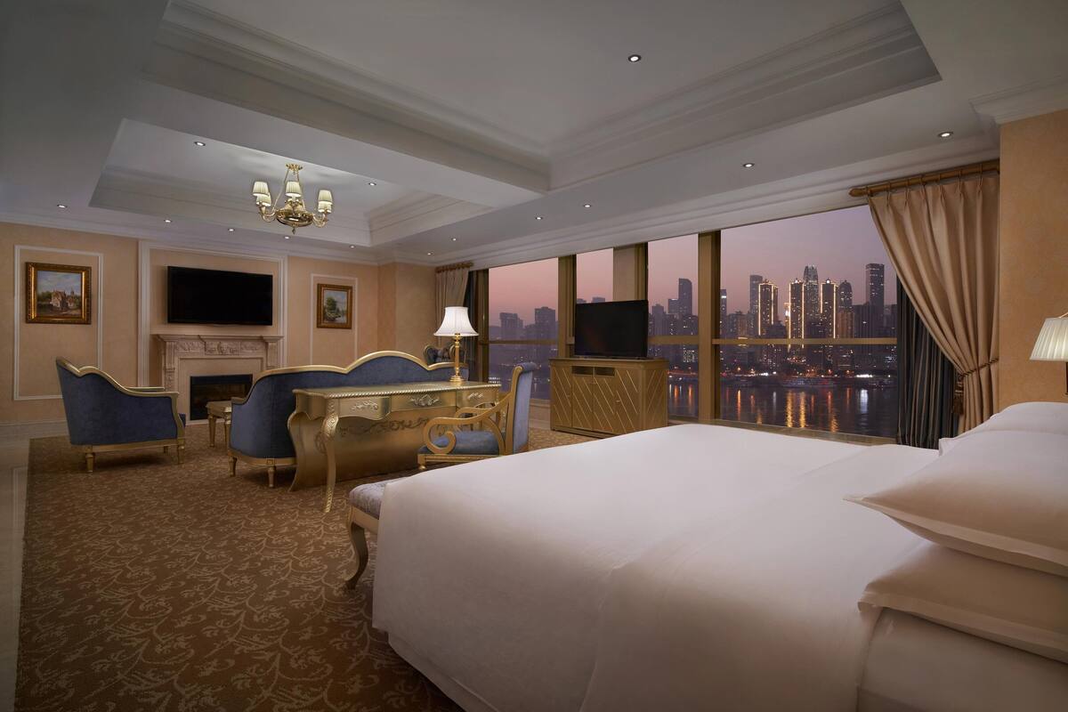 A hotel room with a large bed, a desk with a chair, a television, and a fireplace.