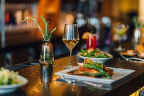 A bar setting with a glass of white wine, a small vase with rosemary, and plates of food.
