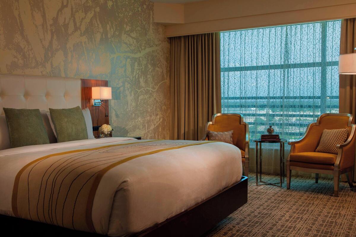 A hotel room with a large bed, a nightstand with a lamp, a chair, and a view of a cityscape through a large window.