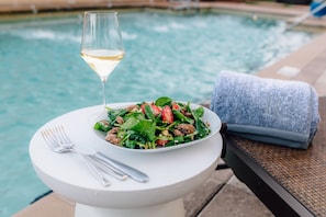 A dining table with a salad, a glass of white wine, and a towel beside a pool.