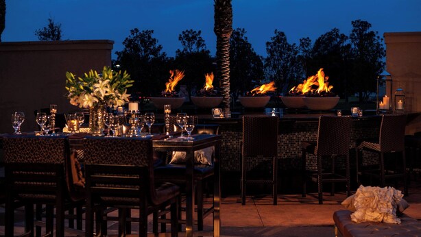 An outdoor dining area with tables set for a meal, surrounded by chairs, and featuring a central fire feature.