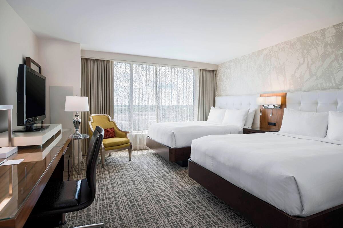 A hotel room with two beds, a desk with a TV, a chair, and a large window with curtains.