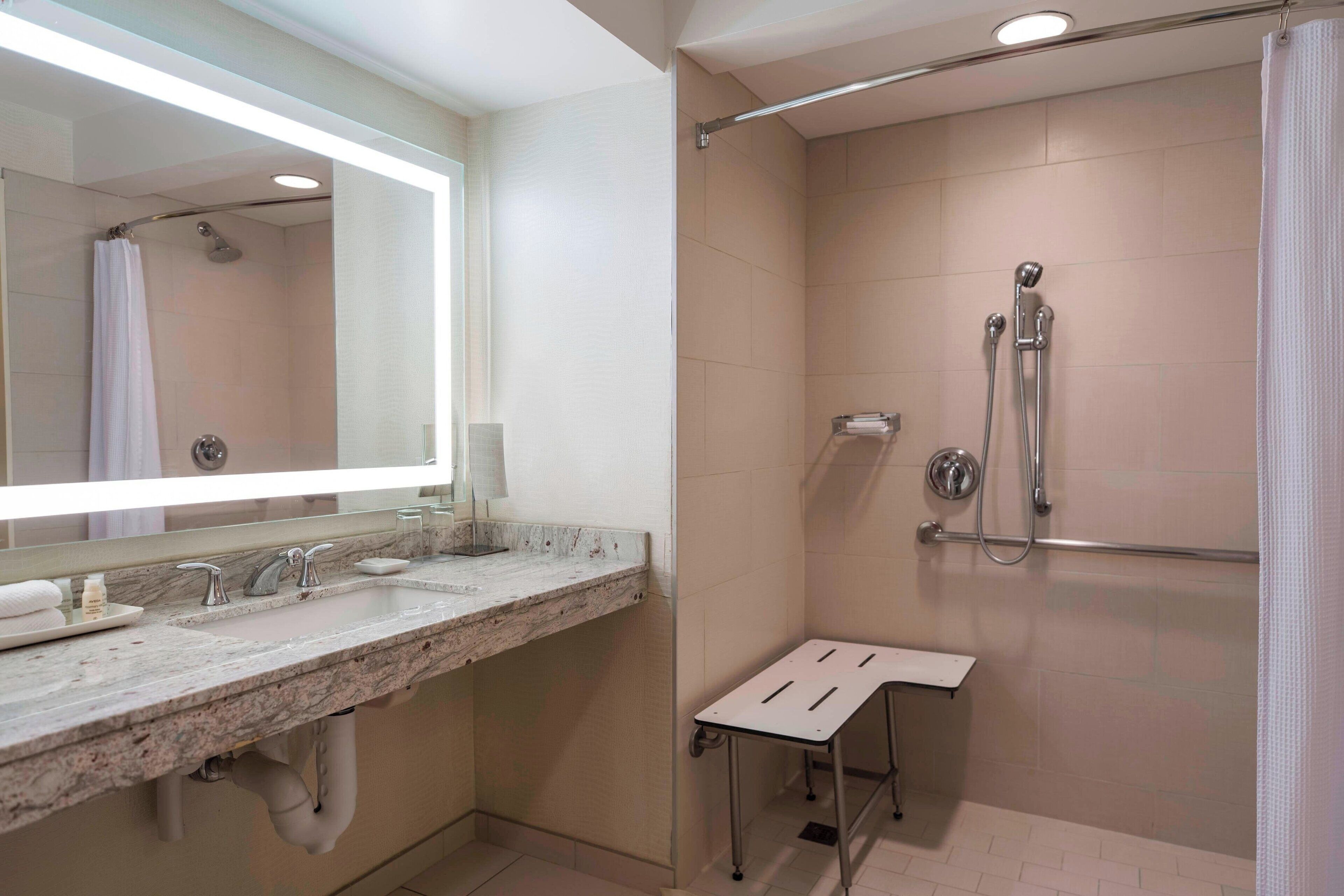 A modern bathroom with a marble sink, a large mirror, a shower with a handheld showerhead, and a small stool.
