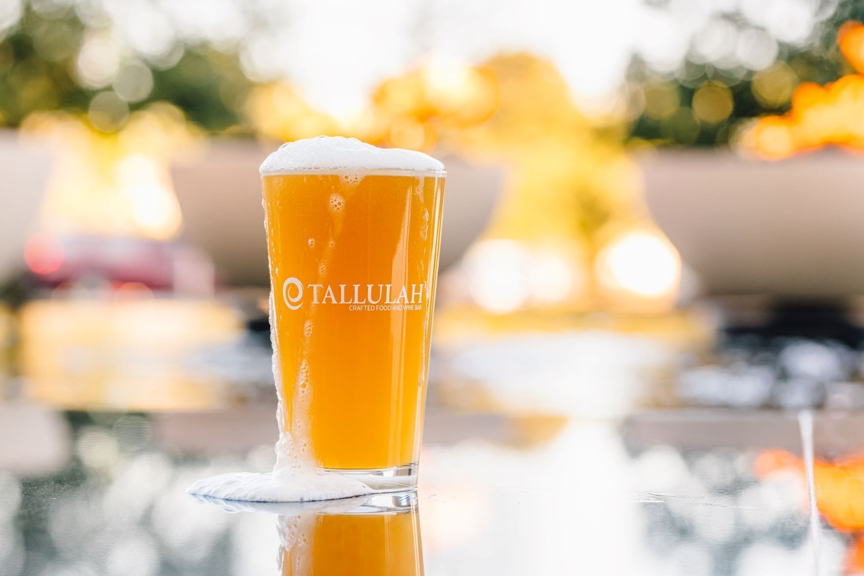 A glass of beer with foam on top, labeled "Tallulah Crafted Food and Wine Bar," on a reflective surface.