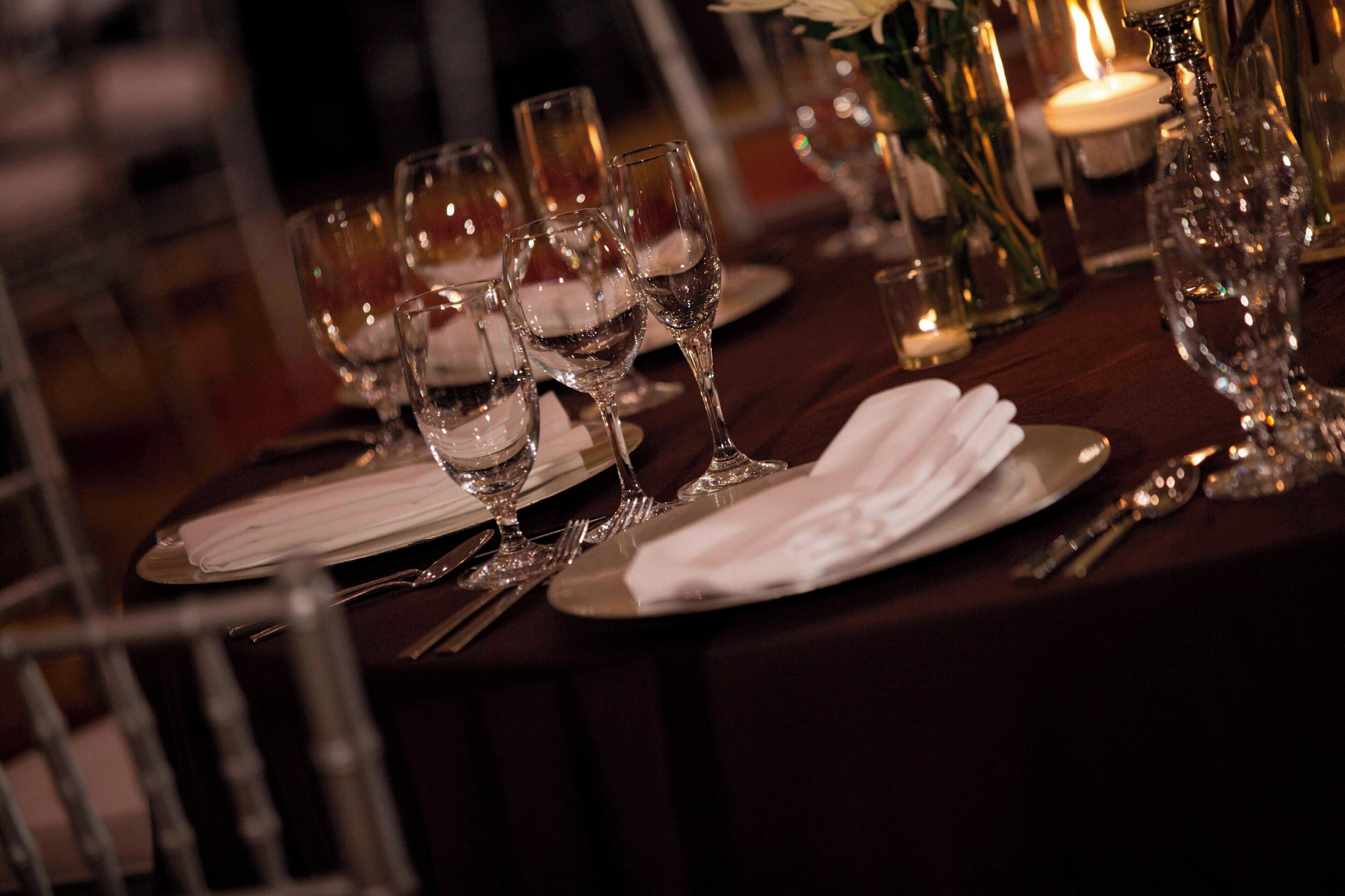 A table set with wine glasses, plates, and cutlery, with a candle and flowers in the background.