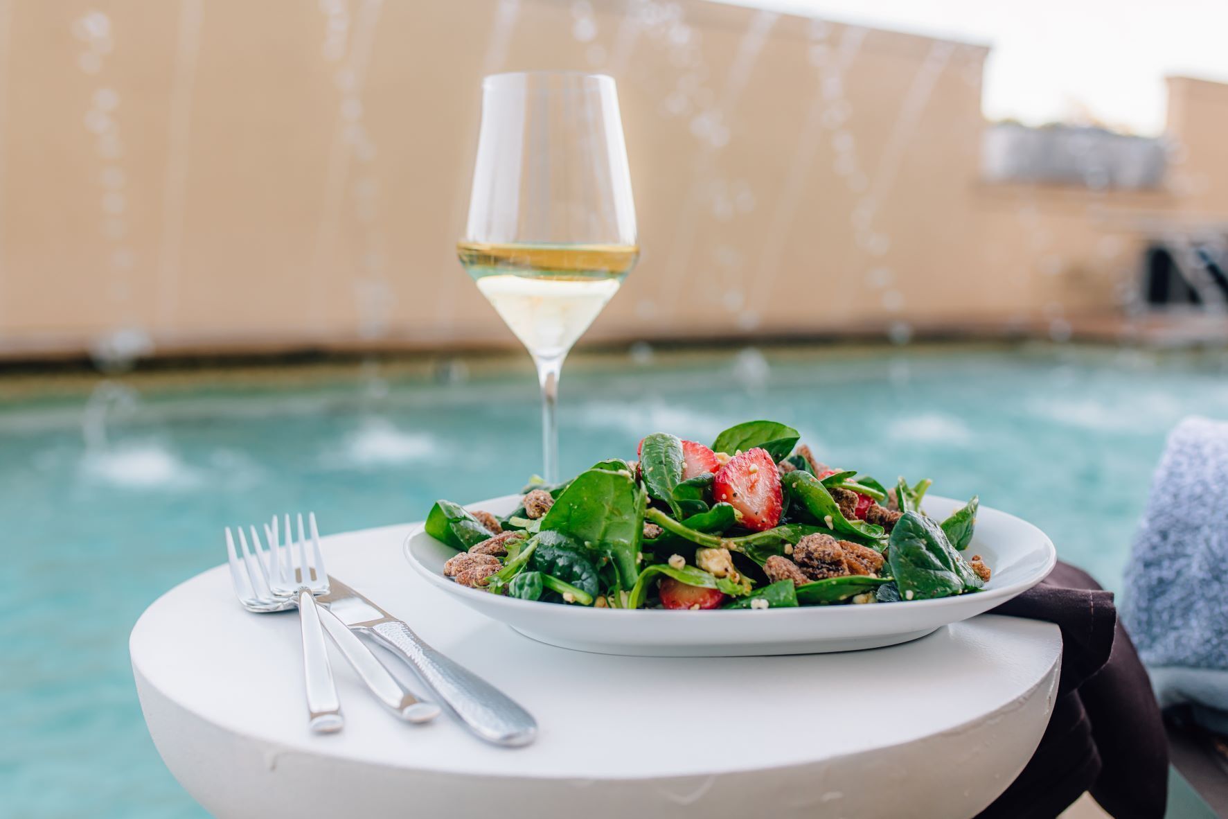 A dining table with a plate of food and a glass of white wine, set against a poolside backdrop.