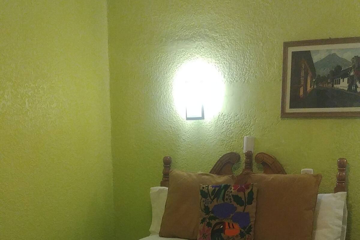Un dormitorio con cama, paredes verdes, una mesita de noche y una lámpara de pared.