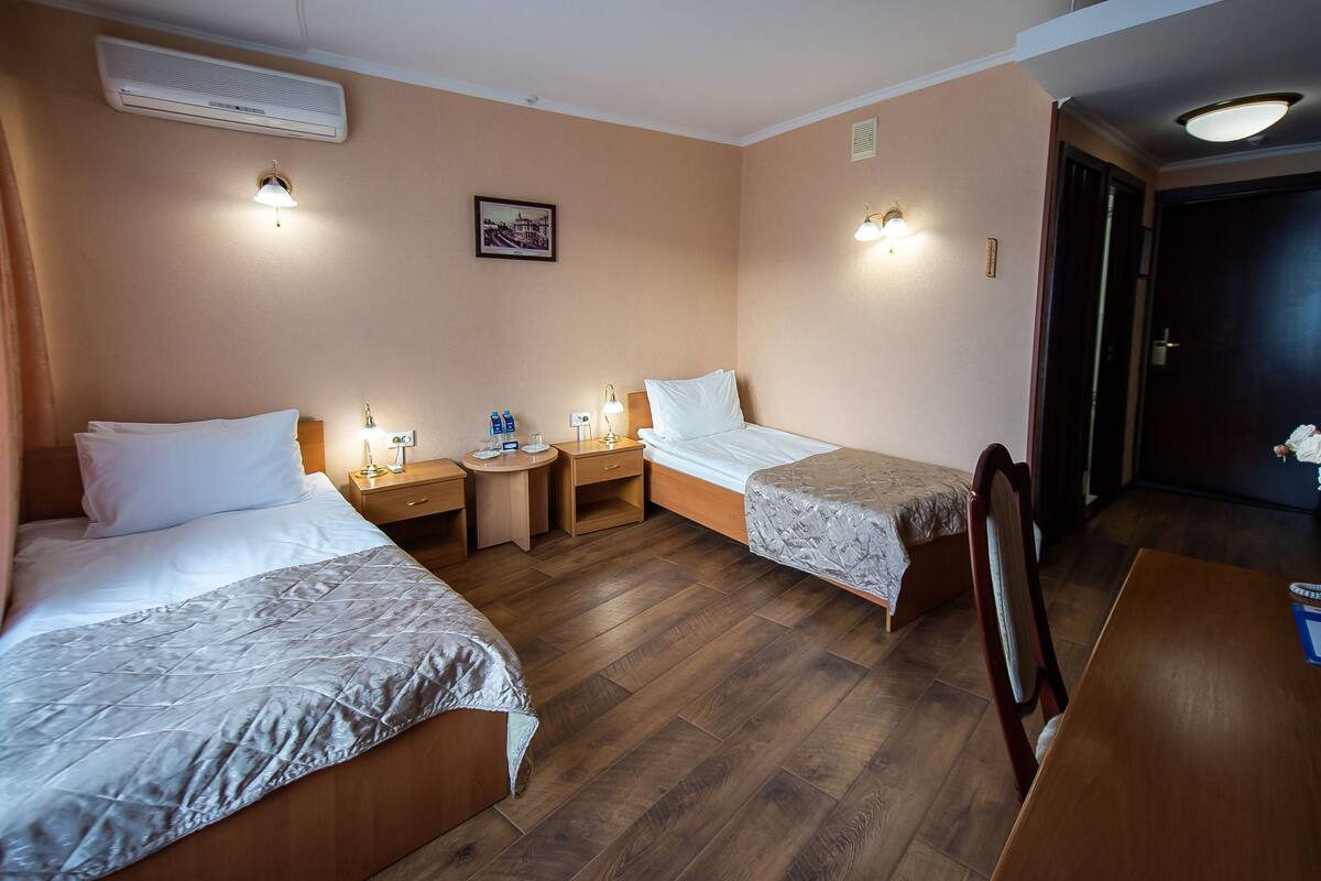 A hotel room with two beds, a wooden floor, a desk with a vase of flowers, and a picture on the wall.