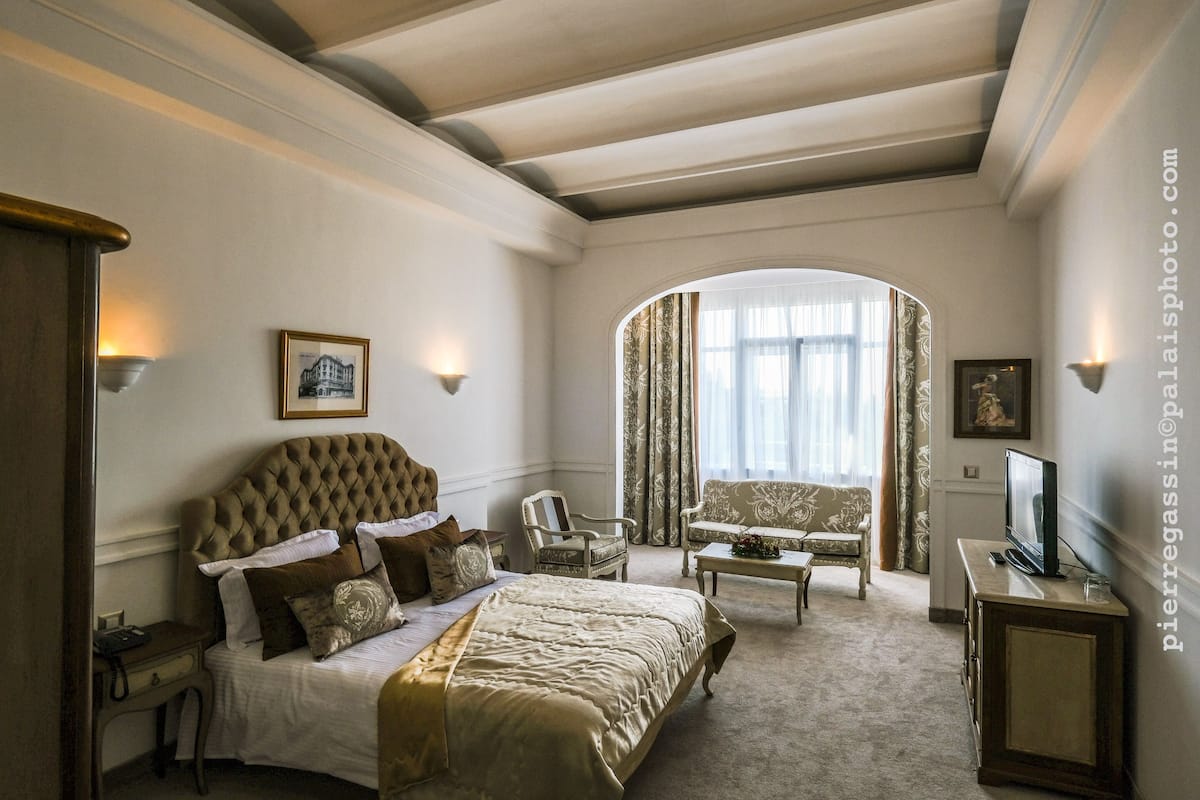 A hotel bedroom with a large bed, a sitting area with a sofa and armchair, a television, and a window with curtains.
