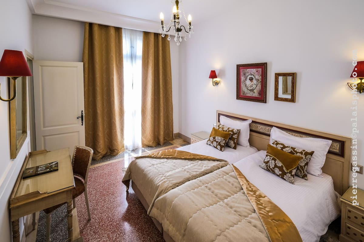 A hotel room with two beds, a desk, a chair, a chandelier, and a door.