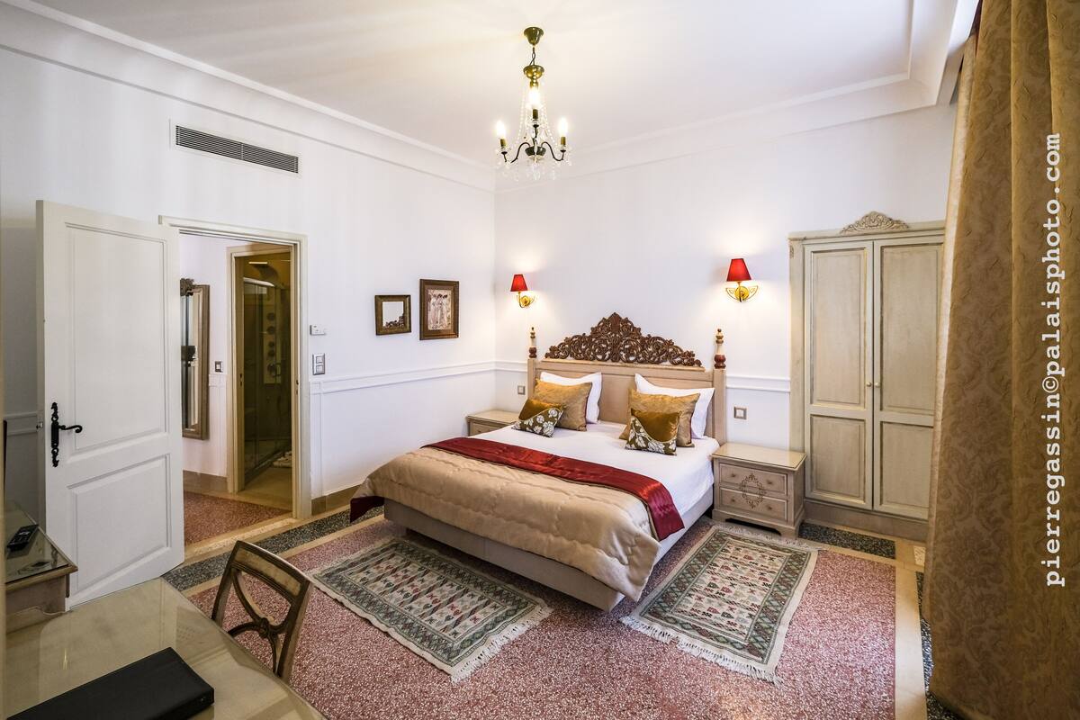 A bedroom with a large bed, two bedside tables, a chandelier, a wardrobe, and a door leading to another room.