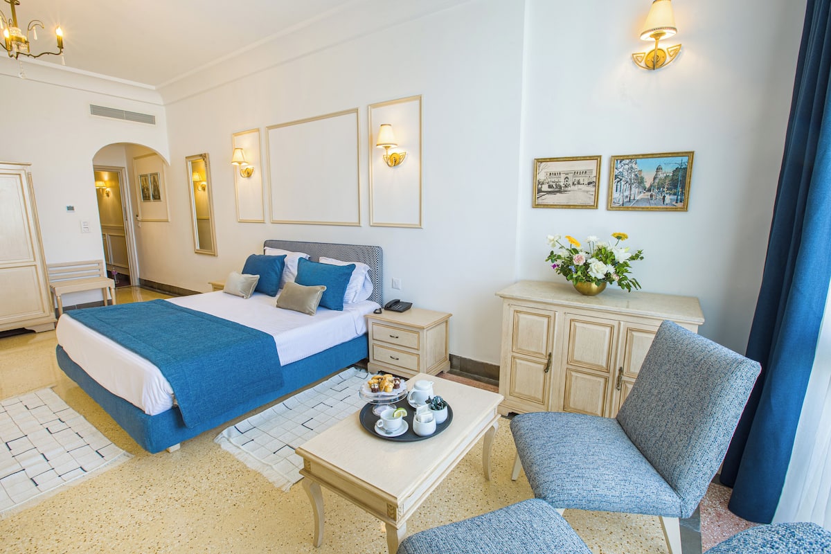 A hotel room with a large bed, bedside table, and a seating area with a chair and table.