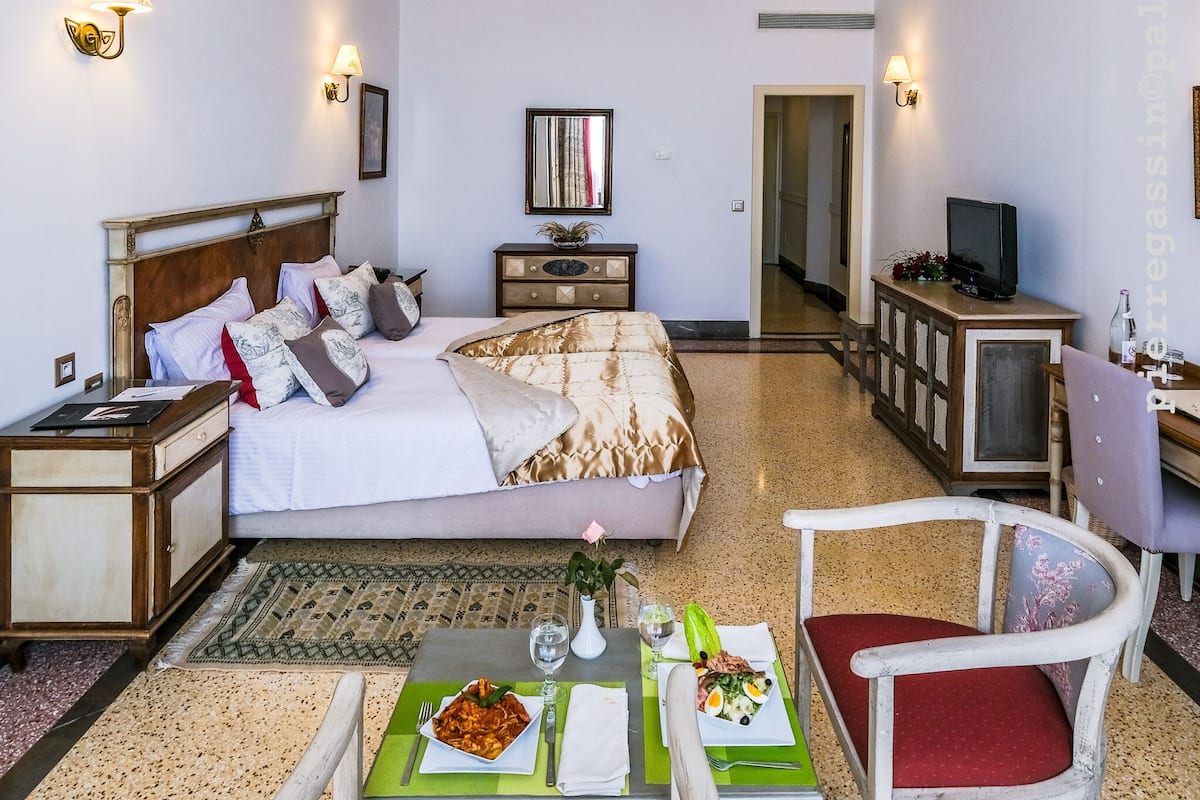 A hotel room with a bed, bedside tables, a TV, a dining area with a table and chairs, and a hallway.