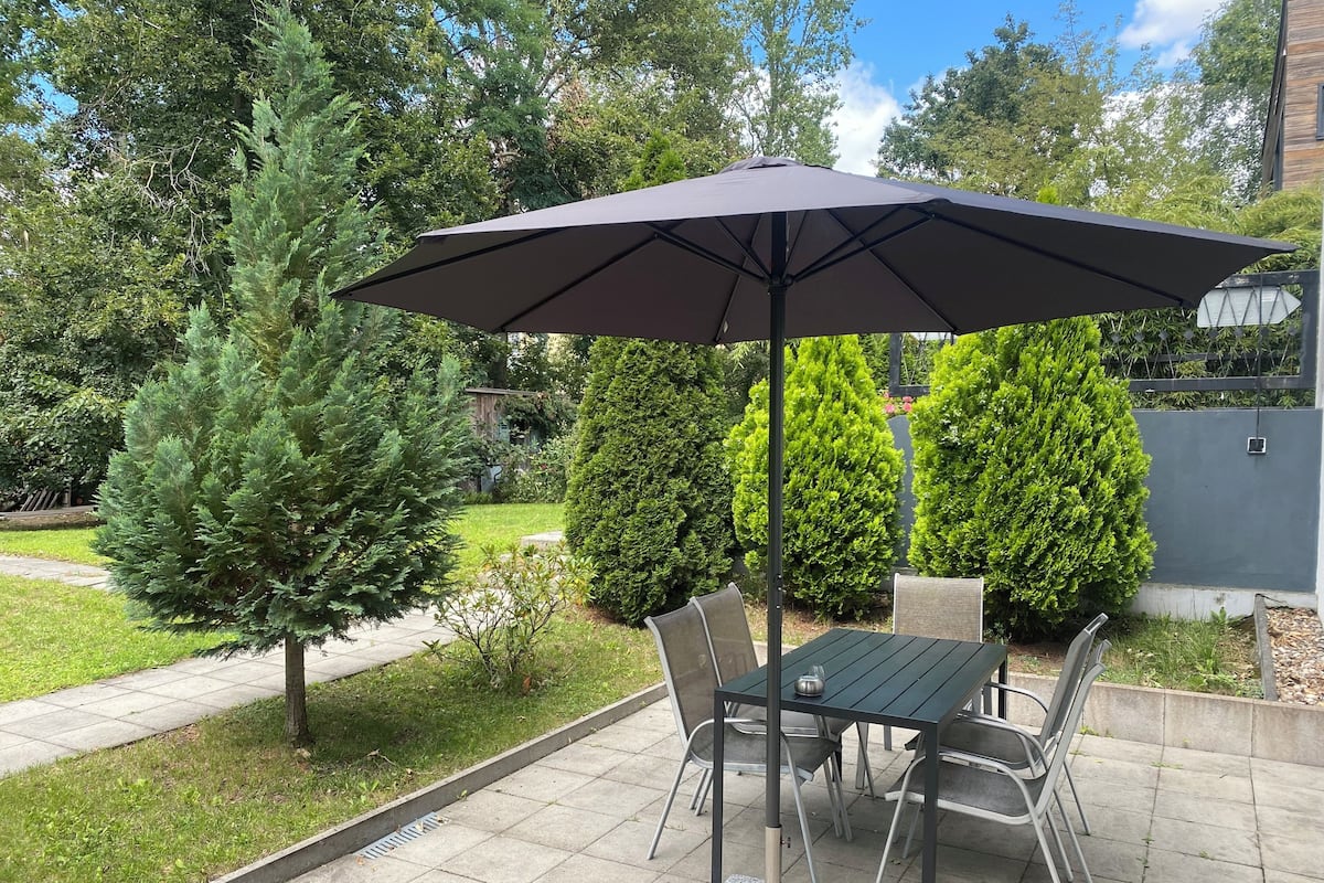 Eine Terrasse im Freien mit Tisch und Stühlen, einem großen Sonnenschirm und einem gepflegten Garten mit verschiedenen Bäumen und Sträuchern.