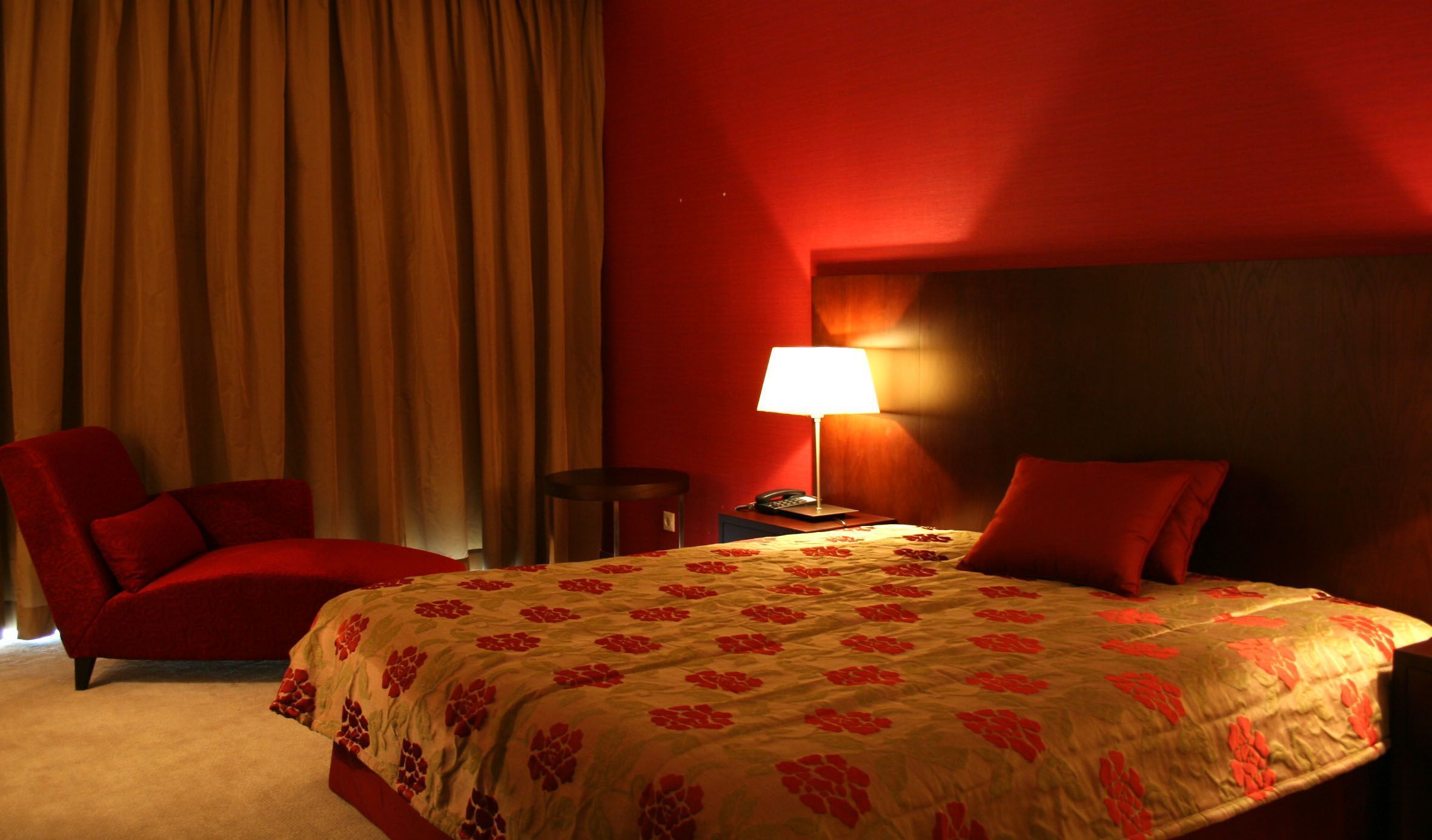 Ein Hotelzimmer mit einem großen Bett, einem roten Sessel, einem Nachttisch mit Lampe und Blick ins Freie durch Vorhänge.