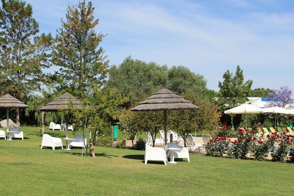 A garden area with outdoor seating, including white lounge chairs and thatched umbrellas, surrounded by trees and a clear sky.