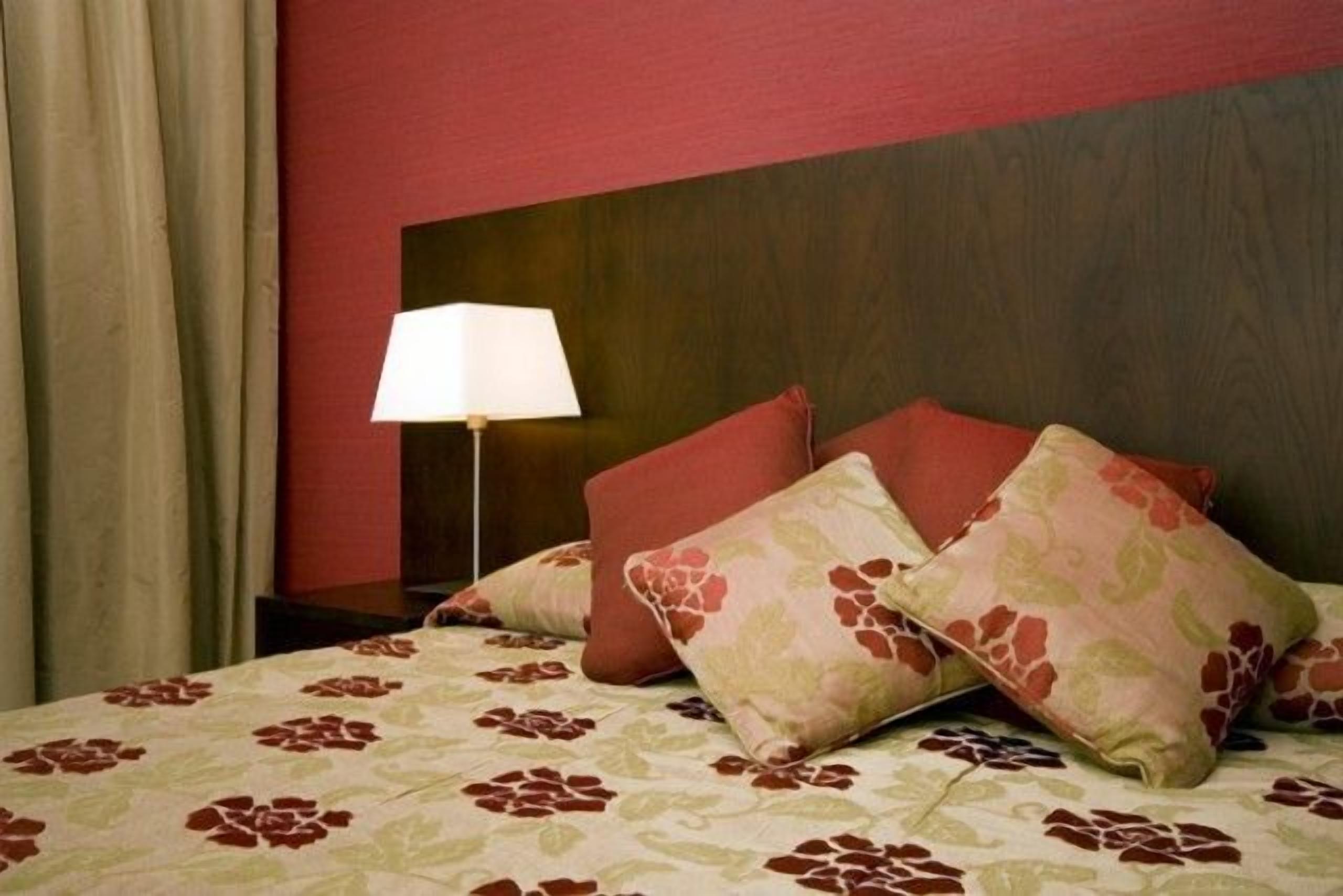 Habitació d'hotel amb llit, calaixera, làmpada i paret vermella.