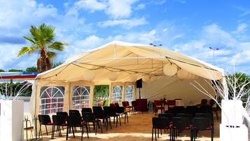 Una carpa grande con múltiples ventanas y un escenario, rodeada de sillas y mesas, ubicada al aire libre.