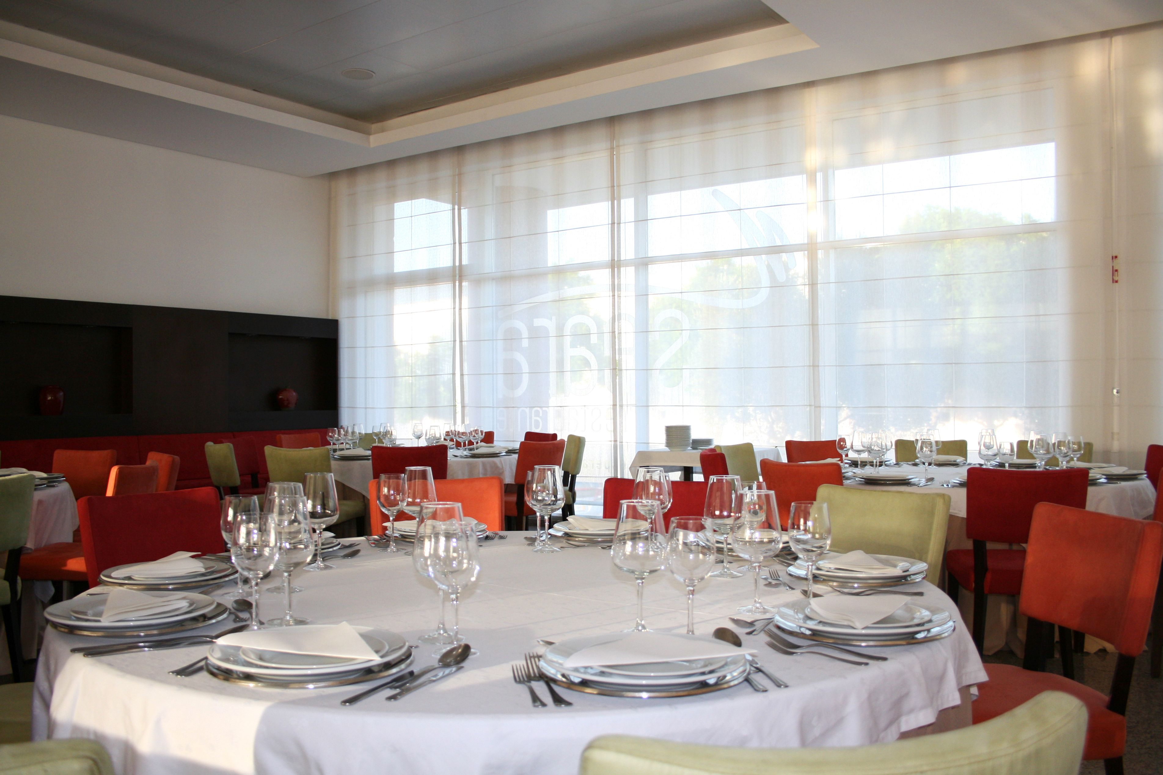 Uma sala de jantar com mesas redondas preparadas para uma refeição, cadeiras vermelhas e amarelas, e janelas amplas com cortinas leves.