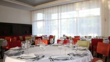 Uma sala de jantar com mesas redondas preparadas para uma refeição, cadeiras vermelhas e amarelas, e janelas amplas com cortinas leves.