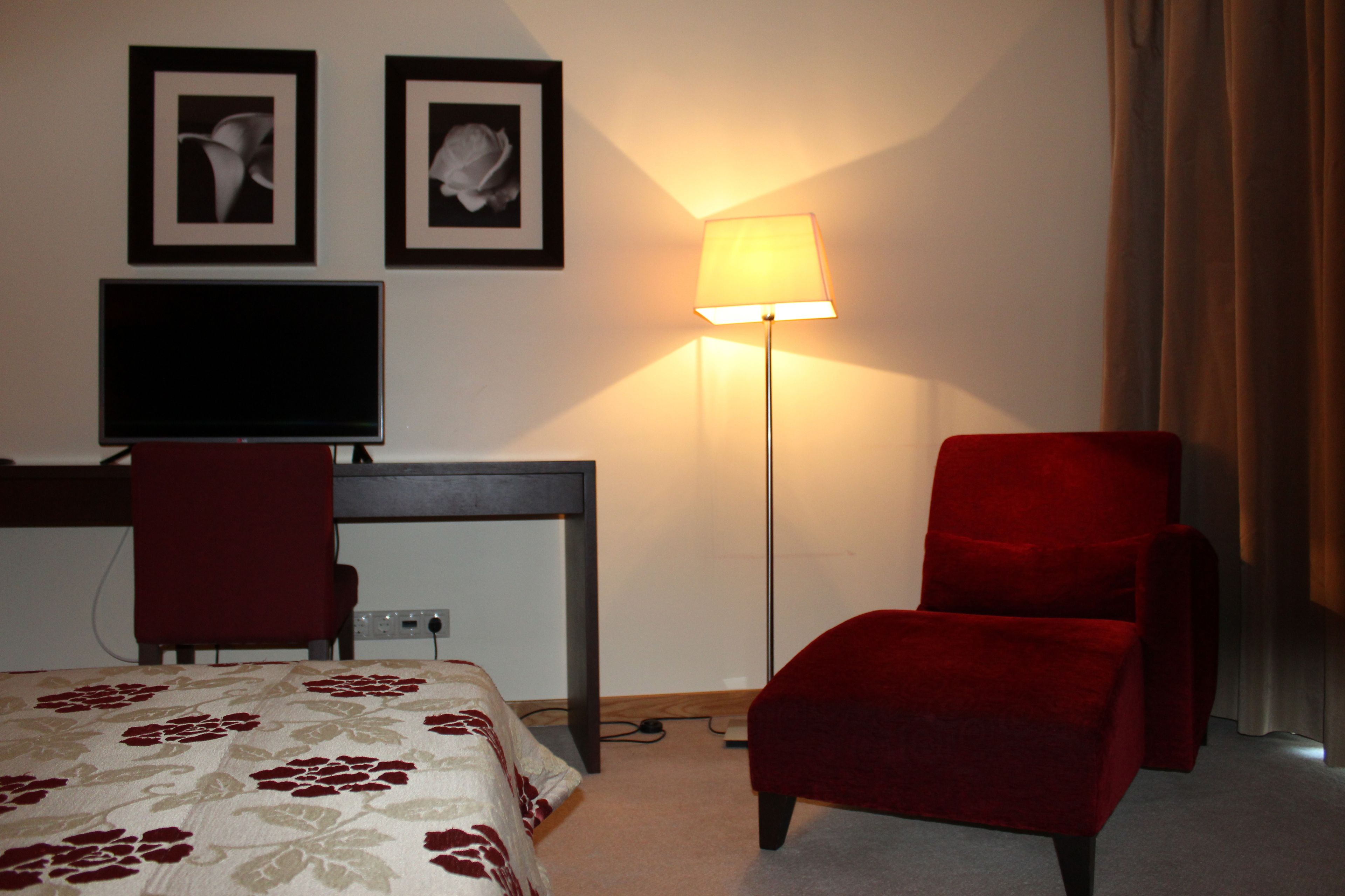 A hotel room with a bed, a desk with a television, a red chair, and a floor lamp.