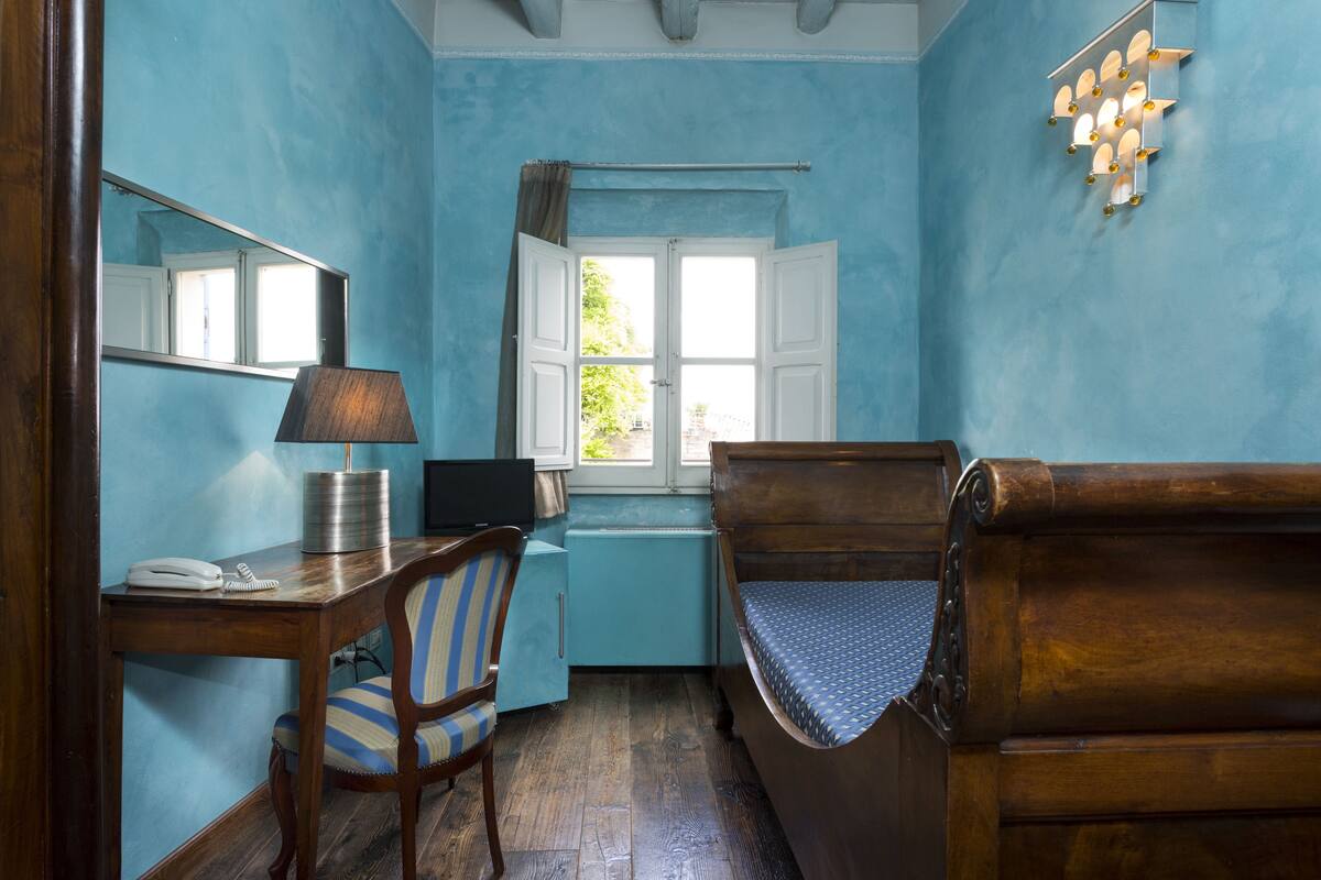 A room with a wooden desk, a chair, a bed, a window with curtains, a mirror, and a wall-mounted light fixture.