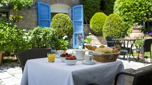 A table set with breakfast items, including bread, fruit, and a jug of orange juice, in an outdoor patio area with a stone wall and blue shutters in the background. - Hotel Il Villino (Santarcangelo di Romagna)