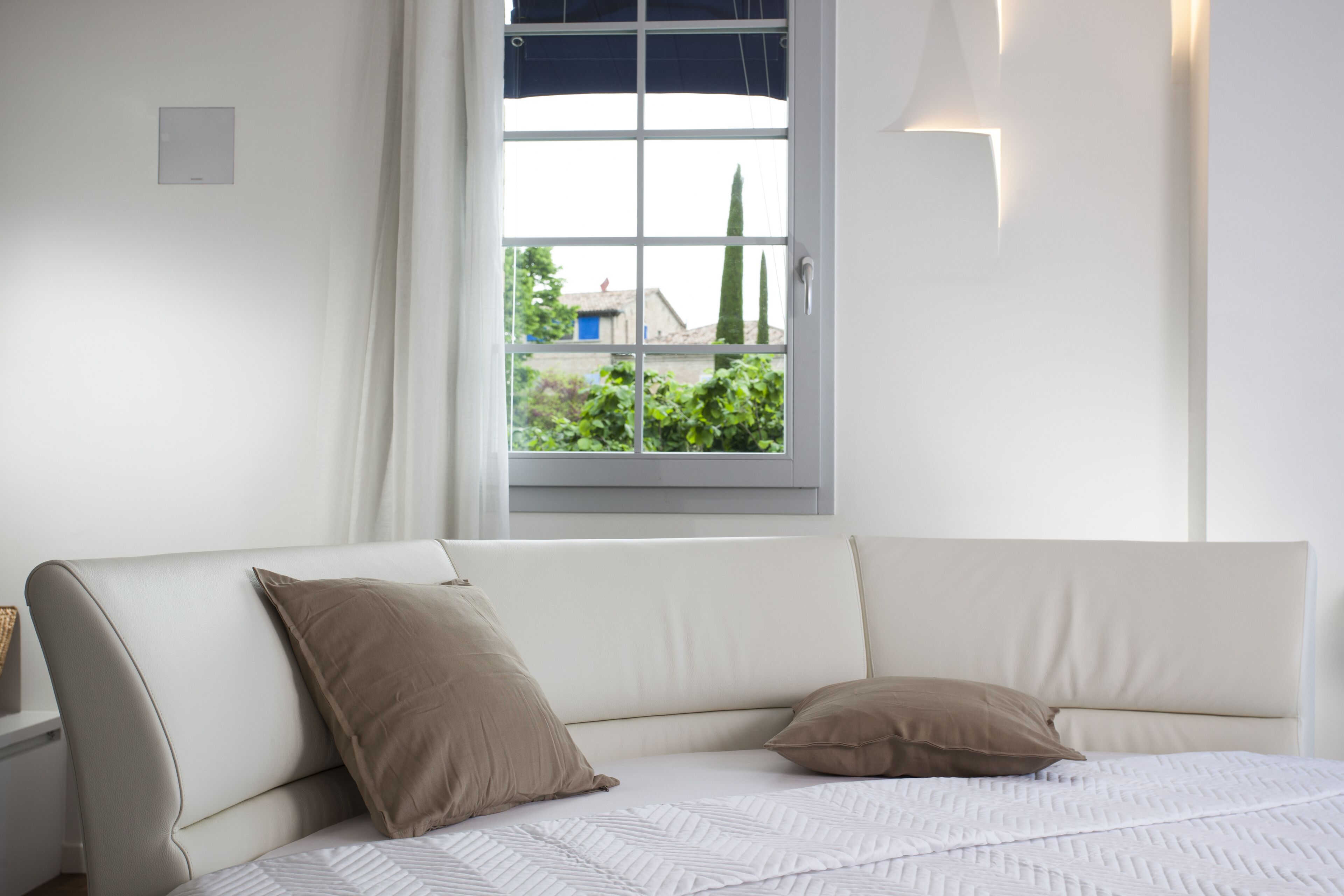 A bedroom with a white bed, two pillows, a nightstand, and a window with a view of greenery and buildings.