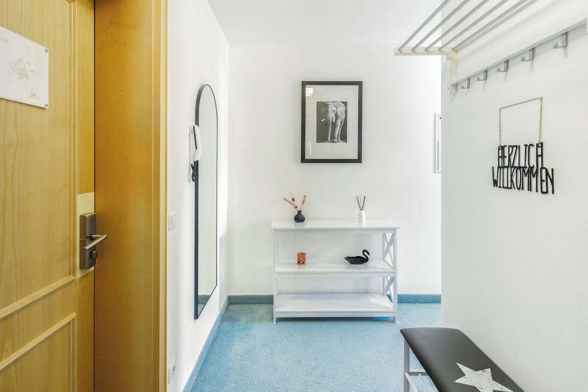 A hallway with a wooden door, a white wall with a framed picture, a white shelf with decorative items, and a bench with a star design.