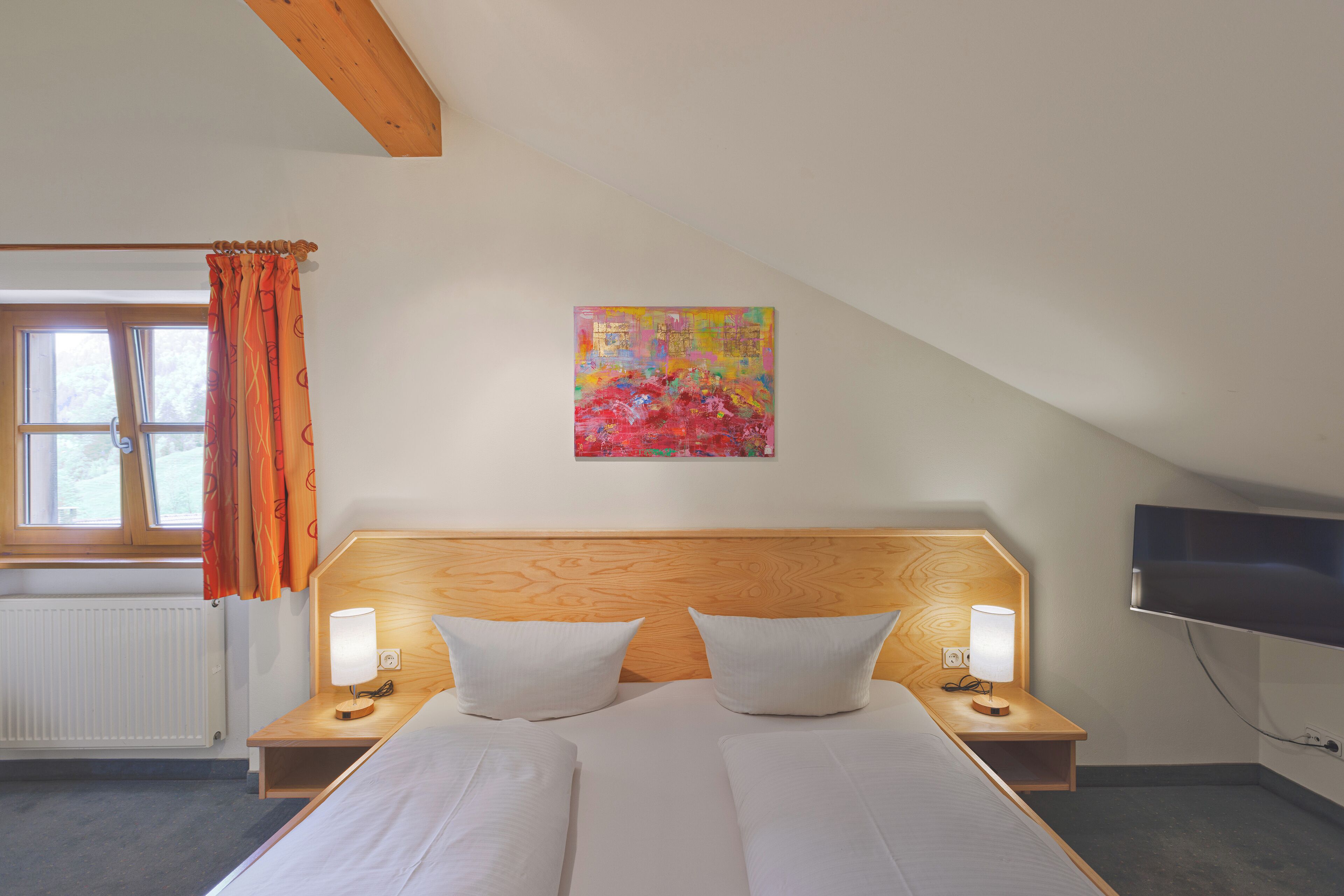 A hotel room with a wooden headboard, two beds, bedside lamps, a TV, and a window with curtains.