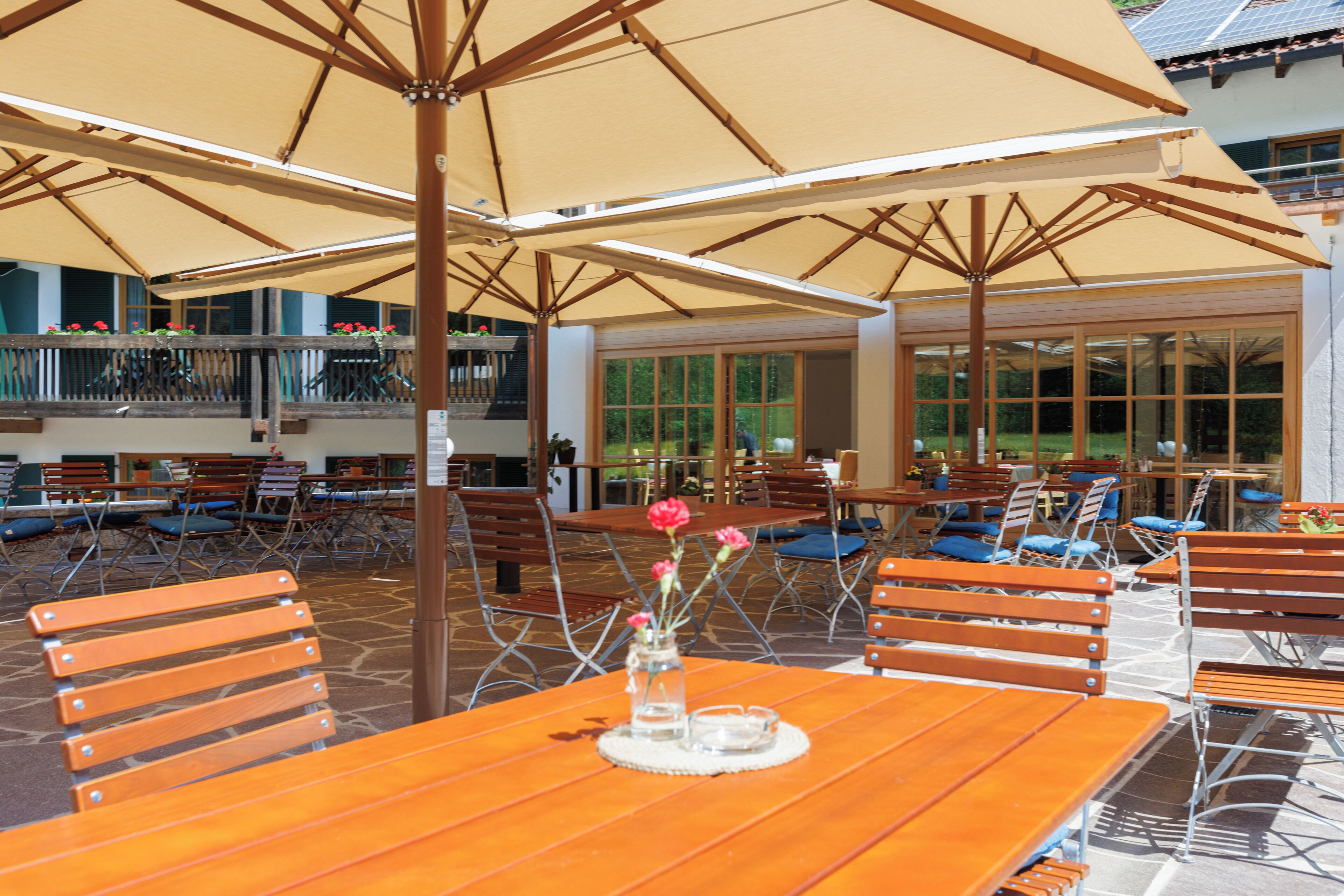 An outdoor seating area with wooden tables and chairs, covered by large umbrellas, and a view of a building with large windows.
