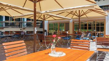 An outdoor seating area with wooden tables and chairs, covered by large umbrellas, and a view of a building with large windows.