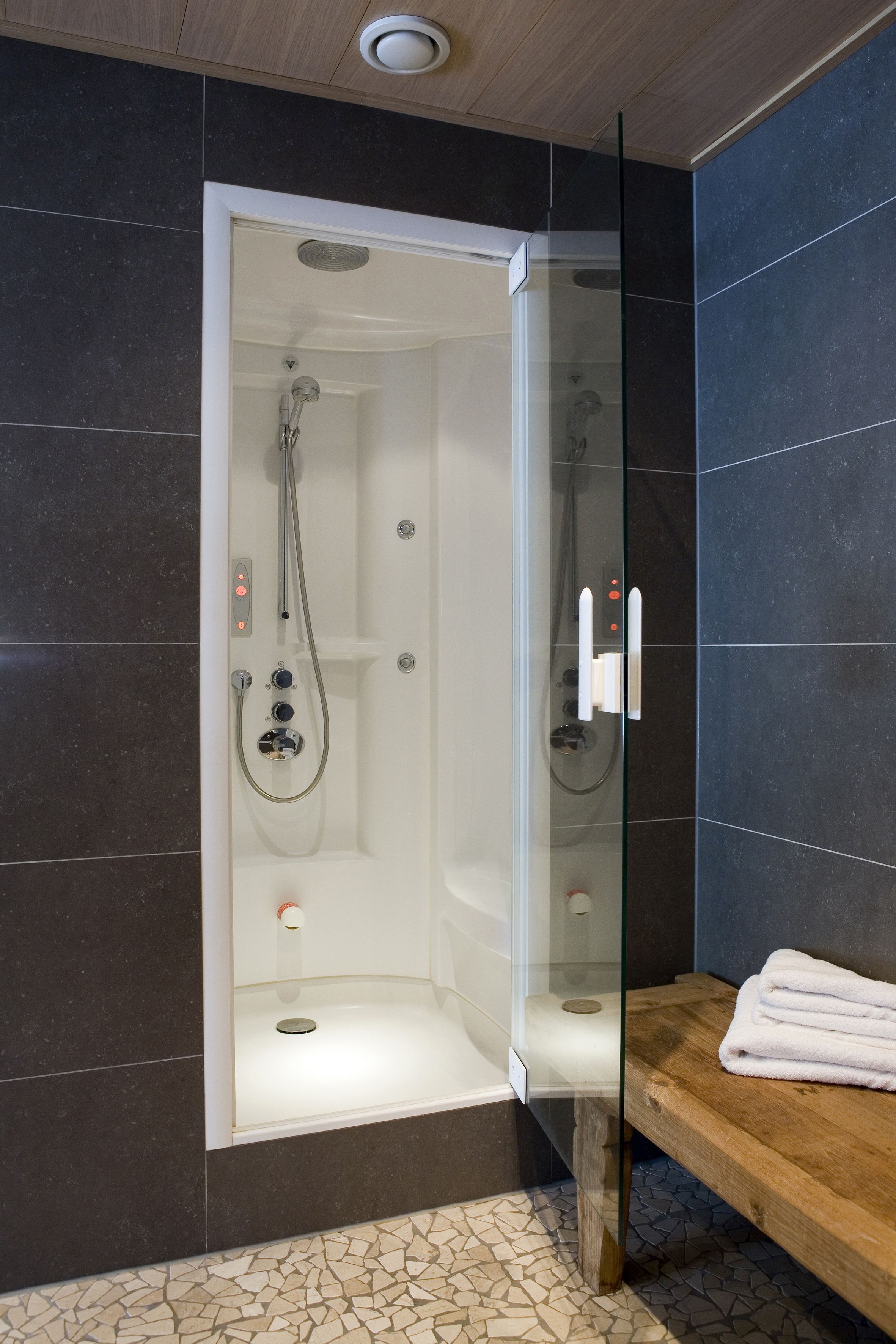 A modern shower enclosure with a glass door, control panel, and a wooden bench with towels.
