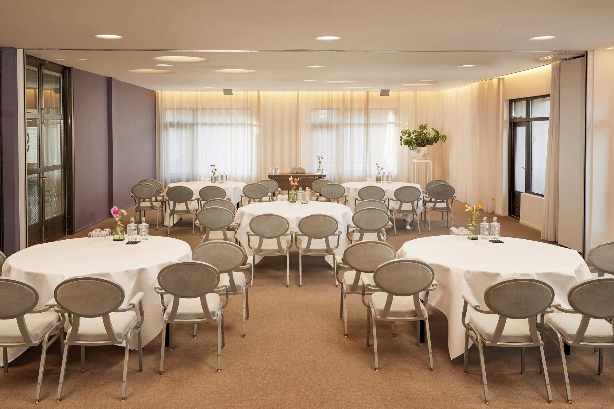 A conference room with round tables and chairs, large windows with sheer curtains, and a central table with floral arrangements.