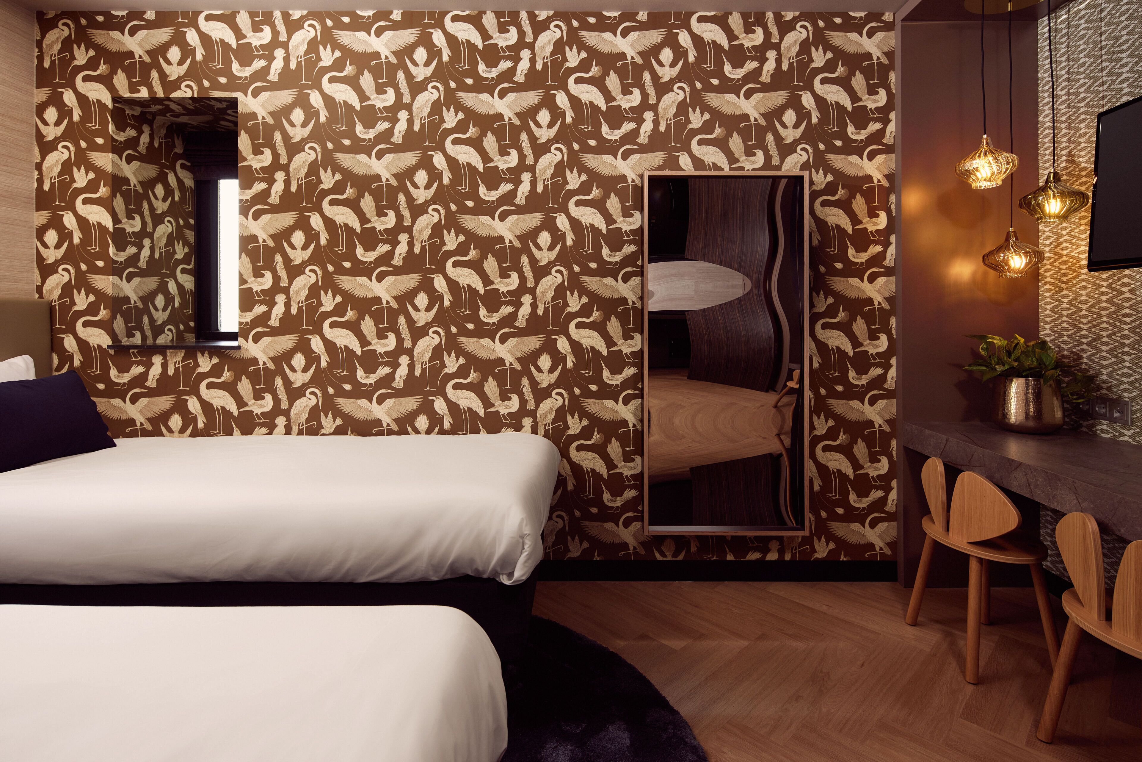 Een slaapkamer met behang met een patroon, een grote spiegel, een bed, een bureau met stoelen en hangende pendelverlichting.