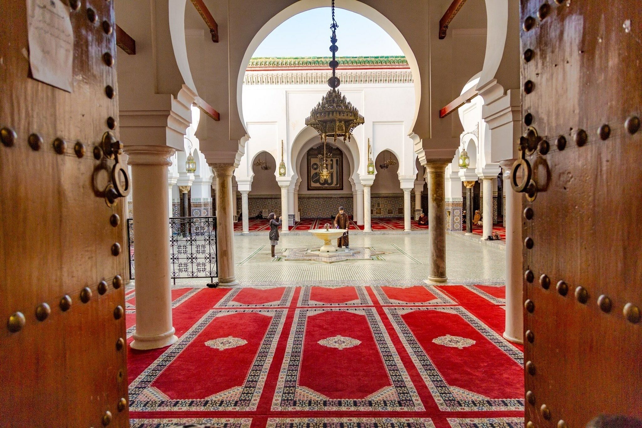 Vue intérieure d’une mosquée avec une fontaine centrale, un tapis rouge à motifs et des colonnes blanches.