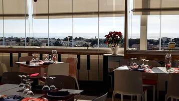 Un espace repas avec des tables préparées pour deux personnes, de grandes fenêtres offrant une vue sur la mer et des luminaires suspendus.