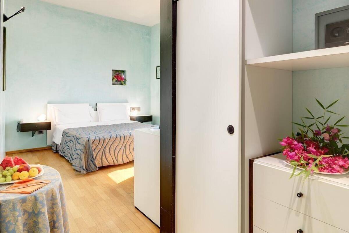 A bedroom with a bed, bedside tables, a small table with fruit, and a wall-mounted shelf with flowers.