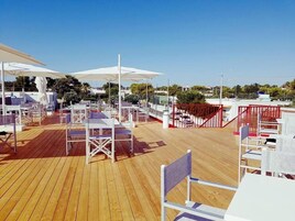 Una terraza al aire libre con piso de madera, mesas y sillas blancas, y grandes sombrillas blancas que dan sombra.