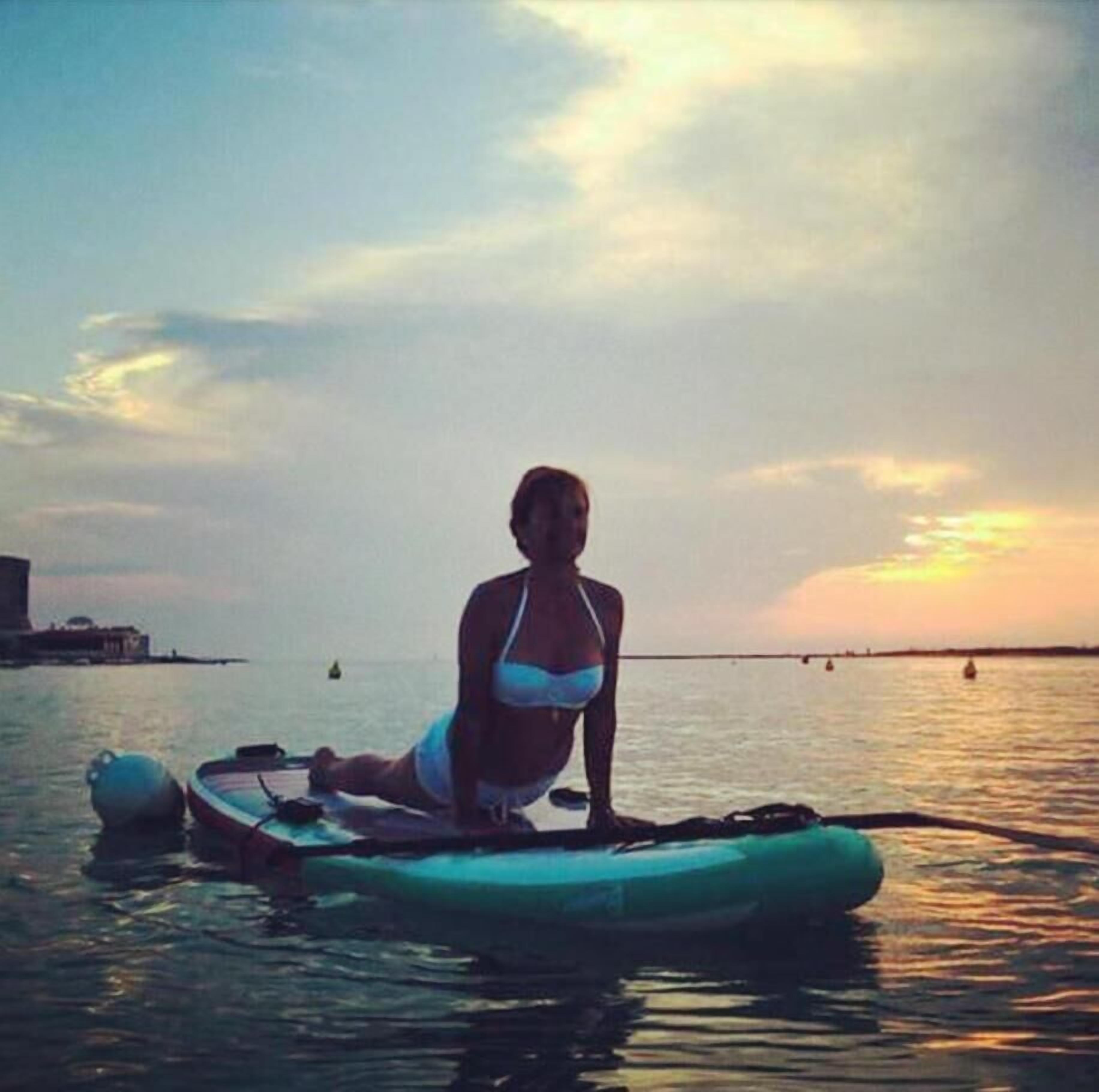 Una persona sentada en una tabla de remo sobre aguas tranquilas, con un atardecer de fondo.